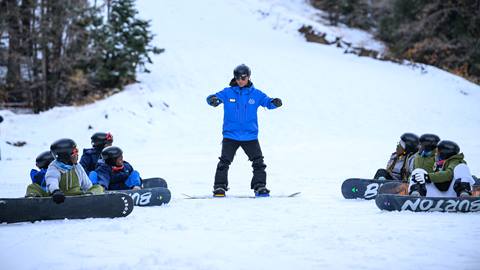 A snowboard instructor in a blue jacket teaching a group of snowboarders who are on the ground starring up.