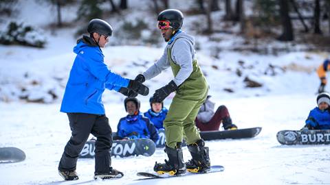 A snowboard instructor teaching a guest on their snowboard with a handful of students in the background on the snow.