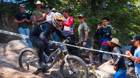A crowd of ten cheering on and watching a mountain biker in all black clothing, full face grey helmet, and grey mountain bike ride down a dirt trail with rocks at Snow Valley.