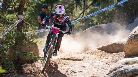 A mountain biker on a teal bike wearing black pants, a black with teal jersey, and white and pink full face helmet riding a dirt trail with trees and rocks surrounding at Snow Valley.