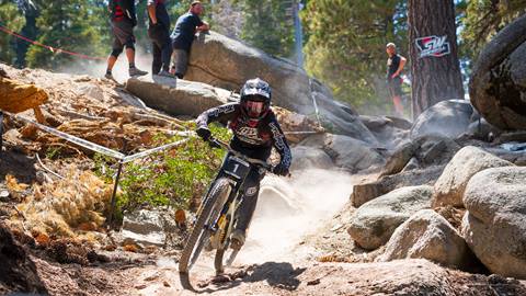 A mountain bike racer in all black attire, black full face helmet, and goggles on their mountain bike rolling through a dirt trail with rocks and event goers watching at Snow Valley.