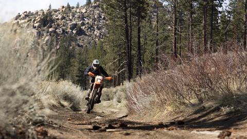 Dirt bike rider on orange bike riding on trail through brush and trees