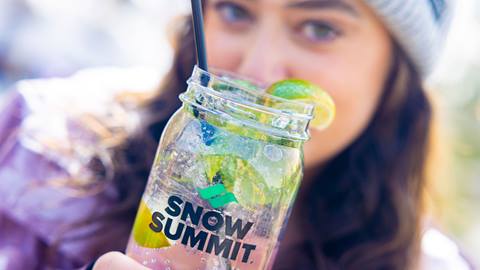 A woman in a purple jacket with grey beanie holding a Snow Summit glass with clear adult beverage in front of her face at Snow Summit.