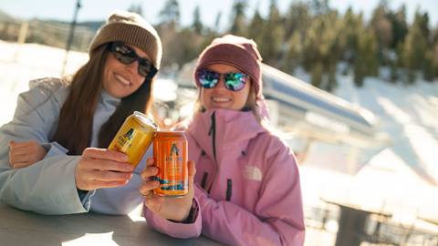 Two adults wearing beanies and sunglasses, one in a blue jacket and the other in a pink jacket, holding Athletic Brewing cans in the base area of Bear Mountain during a sunny winter day.