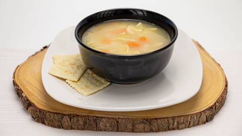 A bowl of chicken noodle soup with crackers on a plate mounted on a wood serving tray.