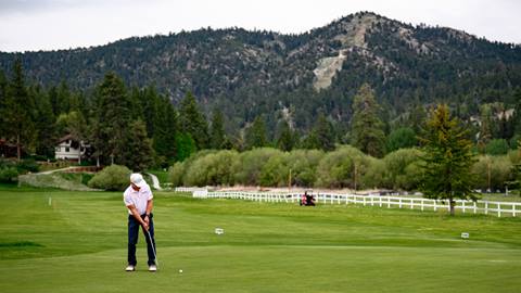 A golfer in a white hat, white collared shirt, and black pants, on a putting green hitting their ball while playing a round at Bear Mountain Golf Course.