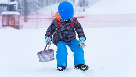 A little kiddo in snow gear holding a easter basket picking up an easter egg in the fresh snow