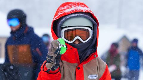 A kiddo in a red and brown Volcom snow jacket, grey helmet, and white goggles holding up a green easter egg
