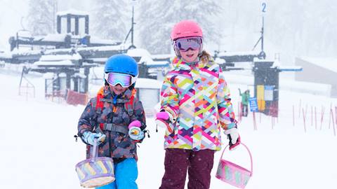 Two kiddos in bright and vibrant snow clothing holding an easter basket in one hand and pink easter eggs in the other hand