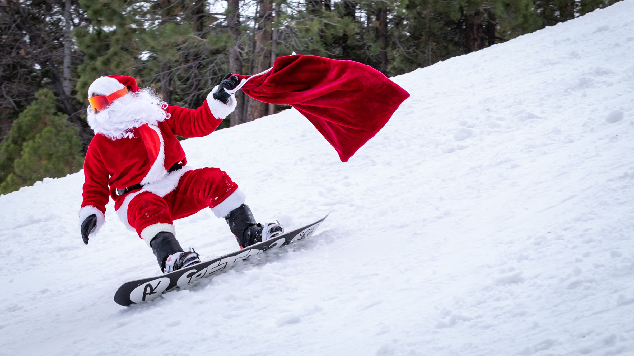 santa snowboarding down a snow ski slope with a red bag in his hands
