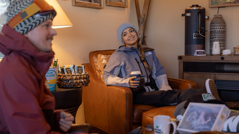 female snowboarder sitting in a chair with a coffee, smiling at her friend in the Ikon Pass Suite at Snow Summit