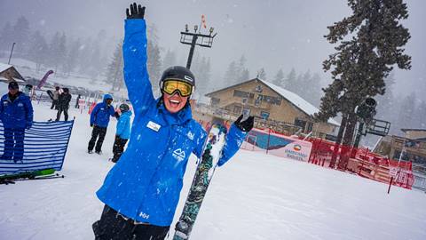 A very excited Snow Valley ski instructor, wearing a blue uniform jacket and goggles with black helmet, with their hands in the air smiling during a wintery storm day.