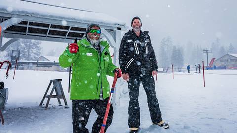 Two winter Snow Valley employees, one wearing all black and the other in black snow pants and a green jacket, standing at the base of a chairlift during a stormy day with snow falling.