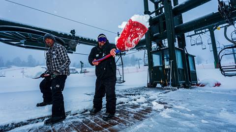 Two Snow Valley winter employees, wearing all black clothes, shoveling snow at the base of the chairlift on a storm day.