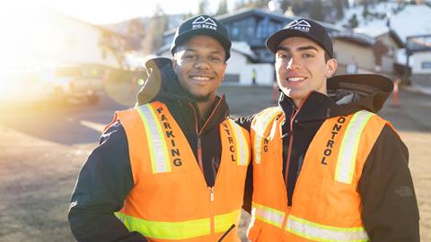 Two parking team members wearing an orange vest, standing in front of the Bear Mountain upper lot parking, smiling at the camera