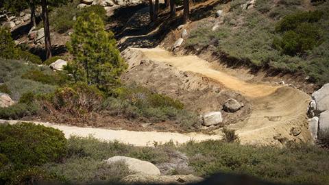 A dirt mountain bike trail at Snow Valley lined with scattered rocks, green bushes, and trees.