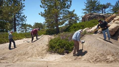 Four trail crew members working on a dirt jump terrain park feature at Snow valley during the day with green trees and shrubs in the background.