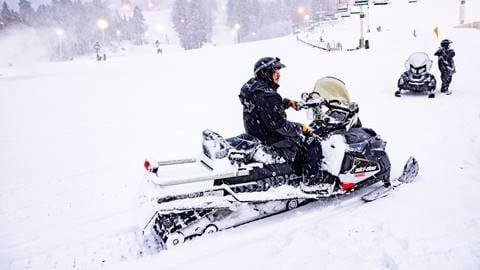 Two Snow Summit employees, one on a snowmobile and the other standing next to one, dressed in all black winter clothing during a snow storm at the base of the hill near a chairlift 