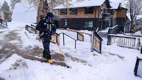 A Big Bear Mountain Resort employee, dressed in their black snow outerwear uniform, wearing black gloves and goggles shoveling snow on the brick walkway in the base area during a snowy day.