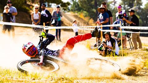 A mountain biker in a full face helmet, black jersey, and red pants who has fallen down on their bike on the dirt in front of a crowd of spectators.