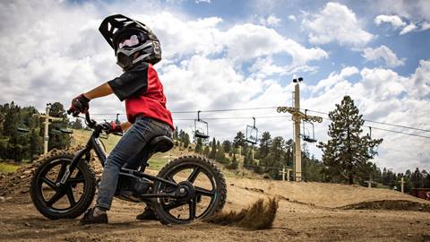 A kiddo on a STAYC electric bike at the base of Snow Summit in a full face helmet with a chairlift and clouds with blue sky in the background.