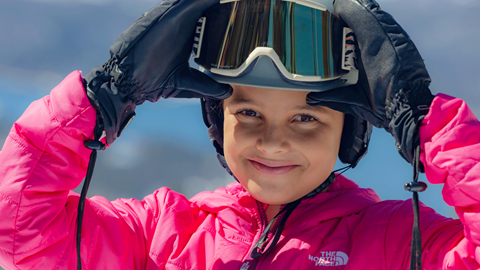 Child wearing pink jacket, holding onto her snow goggles that are on her head