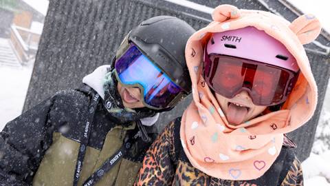 Two kiddos, one in a black and green snow jacket with black helmet and goggles and the other in a colorful leopard snow jacket with pink helmet goggles and a orange head cover with ears, sticking their tongues out to catch the snowflakes during a snowy day.