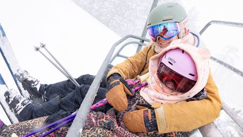 An adult in yellow snow jacket with yellow brown snow mitts and a light color helmet with goggles hugging a kiddo in a colorful leopard snow jacket with pink helmet, goggles and a orange head cover with ears, smiling on a chairlift during a snow day.