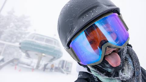 A close up of a kiddo, wearing a black helmet with light lens goggles, in front of a chairlift sticking their tongue out to catch snowflakes on a snowy day.