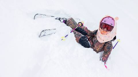 A kiddo in a colorful snow outfit with pink helmet and rose lens goggles sitting in the snow on their skis during a snowy day.