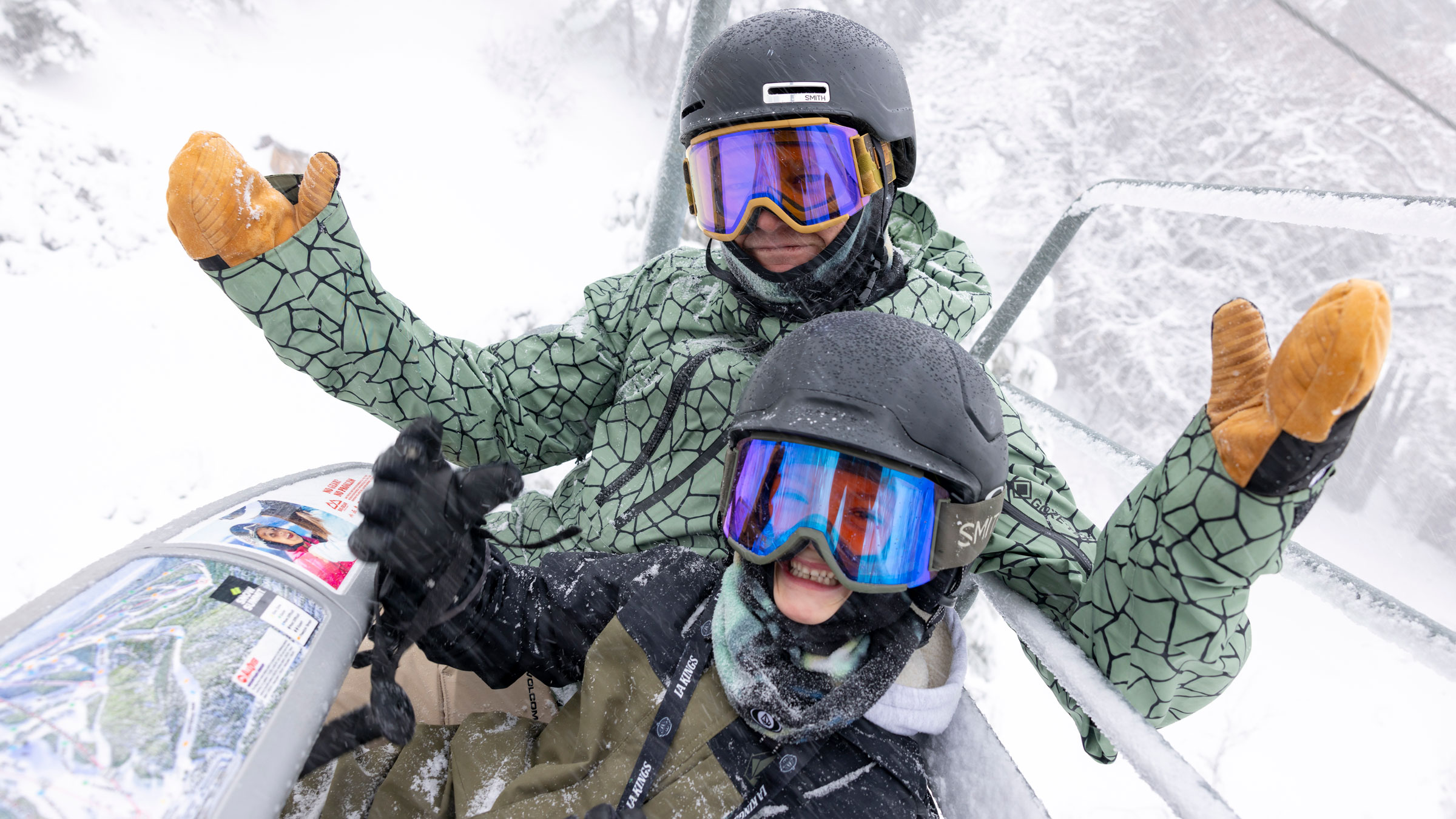 An adult in a green snow jacket with black helmet and yellow snow mitts riding on a chairlift with the safety bar down with their kiddo wearing a green and black jacket with black helmet and goggles during a snow day.