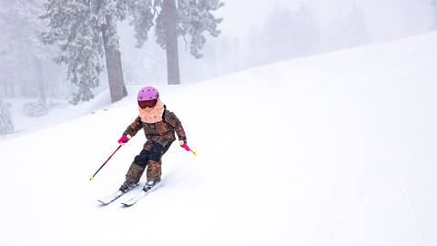 A kiddo in a colorful snow outfit with pink helmet and rose lens goggles skiing down a snow covered trail during a snow storm.