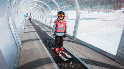 A child on skis at Snow Valley, wearing black snow pants with a pink lesson bib and black helmet, taking a ride on the moving carpet that transports them from the base area to the top of the beginner trail during lessons.