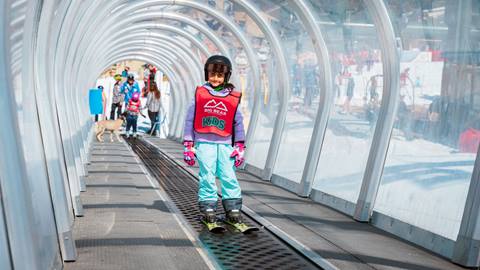 A kid on skis at Snow Valley, wearing teal snow pants with a pink lesson bib and black helmet, taking a ride on the moving carpet that transports them from the base area to the top of the beginner trail during lessons.