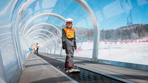 A kiddo on their snowboard at Snow Valley, taking a ride on the moving carpet that transports them from the base area to the top of the beginner trail during lessons.