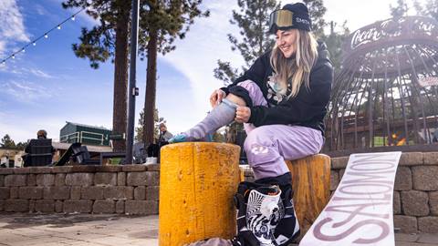 An adult snowboarder in black jacket, black goggles, and light snow pants putting on grey and teal Darn Tough winter socks at the base area of Snow Summit sitting next to a Coca-Cola firepit
