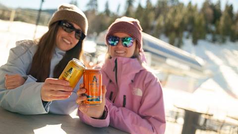 Two adults, one in a pink jacket with beanie and the other in a light blue jacket with beanie, holding cans of Athletic Brewing beverages in their hand during a sunny winter day.