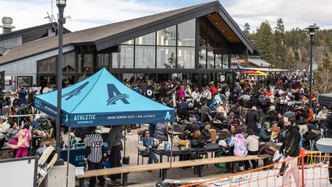 Bear Mountain during winter with skiers and snowboarders sitting on the sun deck in front of Laybacks Bar with a blue Athletic Brewing activation tent in the left corner.