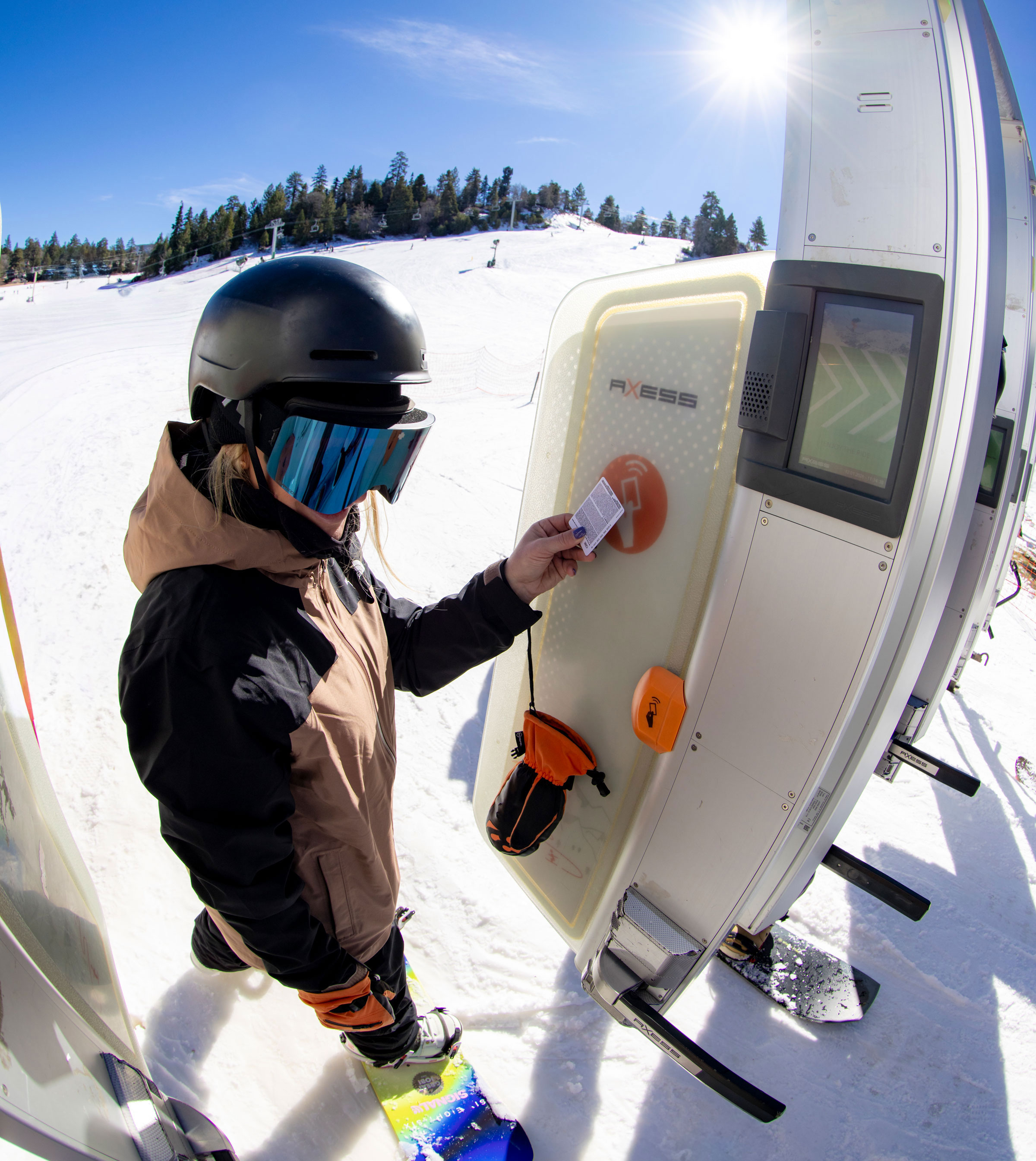 An adult in a brown and black winter jacket, black goggles, and a black helmet scanning their season pass at a lift gate at the base area of Bear Mountain on a sunny winter day.