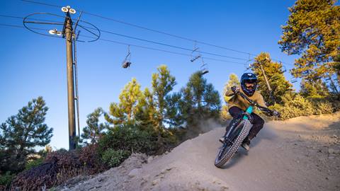 A mountain biker in black pants, yellow long sleeve shirt, and full face helmet riding a teal mountain bike through a dirt berm at Snow Valley.