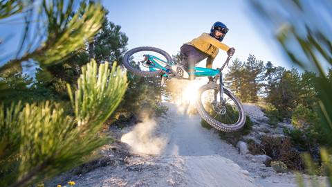 A mountain biker in black pants, yellow long sleeve shirt, and full face helmet riding a teal mountain bike catching air off a Snow Valley bike park dirt jump.