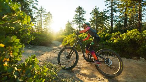 A mountain biker, in a matching Fox branded top and bottom mountain bike apparel set with a full face helmet on a black mountain bike, riding along a dirt trail as the sun sets during summer.