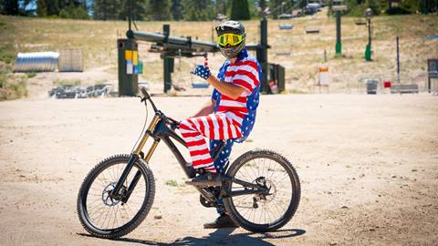 A mountain biker at the base of Snow Valley sitting on their bike in a full face helmet, goggles, and a matching american flag jersey plus pant set.