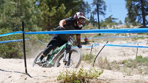 A mountain bike rider, wearing black shorts and black shirt with full face helmet and glasses, riding on a green bike on a loose dirt trail at Snow Valley during a competition.