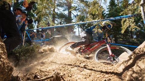 A mountain bike rider in a full face helmet on a red bike racing on a dirt trail with spectators cheering behind a blue roped off area.