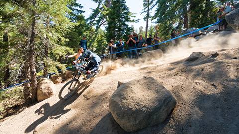 A mountain bike rider in a full face helmet, riding on a dirt trail with large boulders and a crowd cheering from the sidelines.