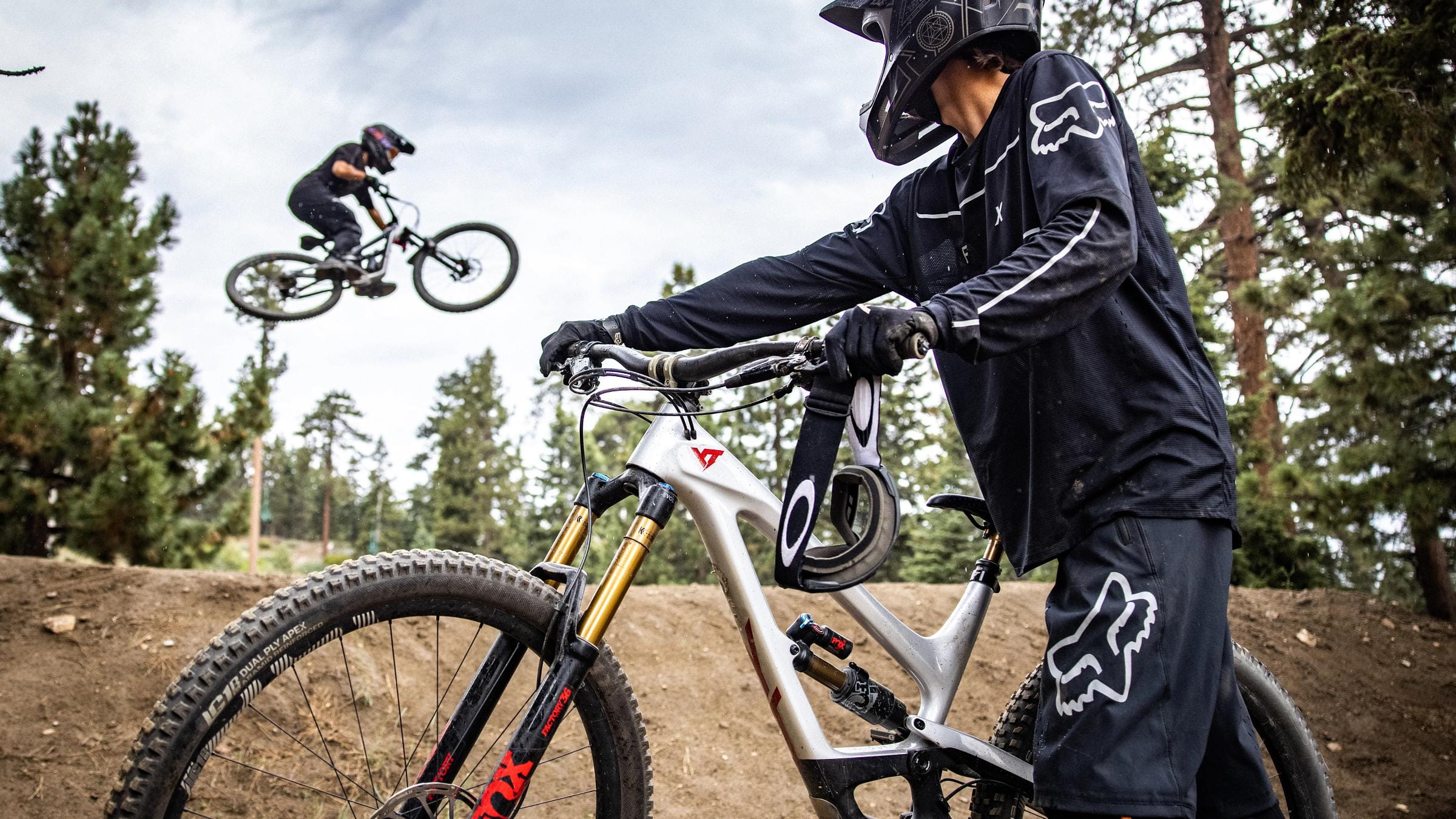 One mountain biker watching the other mountain biker do a jump