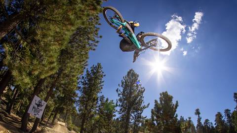 A mountain biker catching air off a dirt jump riding a teal mountain bike at Snow Summit bike park.