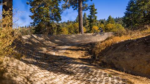 A solo mountain biker in black pants, black long sleeve shirt, and full face helmet riding through a pavement berm on top of dirt with trees surrounding at Snow Summit's Bike Park