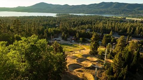 A drone image at sunset of a dirt snake run bike trail at Summit Bike Park with forest views and Big Bear Lake in the background.